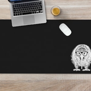 May include: Black desk mat with a white illustration of a kiwi bird wearing glasses. The mat is on a light wood surface.