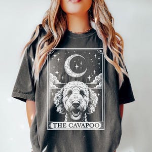 May include: A dark gray t-shirt with a white graphic of a dog with a crescent moon and stars. The text "THE CAVAPOO" is printed below the graphic.