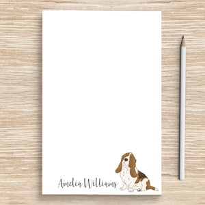 May include: A white notepad with a brown and white basset hound illustration and the name "Amelia Williams" printed at the bottom. A white pencil is lying next to the notepad on a wood surface.