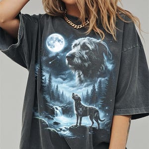 May include: Dark grey t-shirt with a wolf howling at the moon design. The graphic includes a waterfall, forest, and a large wolf head. The shirt has short sleeves and a relaxed fit.
