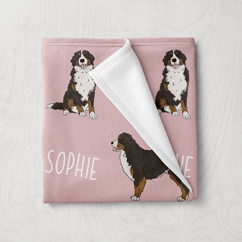 Puede incluir: Una manta de forro polar rosa con un reverso de forro polar blanco. La manta presenta un patr&oacute;n repetido de un perro de monta&ntilde;a bern&eacute;s con el nombre "Sophie" impreso en la manta.