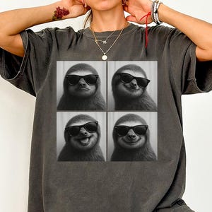 Peut inclure: T-shirt gris foncé avec une image en quatre panneaux d'un paresseux portant des lunettes de soleil. Le paresseux affiche différentes expressions, dont un rictus et un sourire. Le t-shirt est de coupe décontractée.
