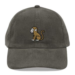 May include: A dark green corduroy baseball cap with a small embroidered monkey design in brown and tan. The cap has a curved brim and a structured crown, suitable for casual wear.