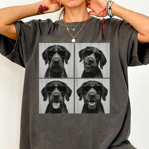 May include: Dark grey t-shirt with a four-panel image of a dog wearing sunglasses. The dog has a black coat and is shown in different poses, including smiling and with its tongue out. The shirt has a relaxed fit.