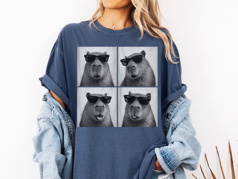 Puede incluir: Camiseta azul marino con una imagen de cuatro paneles de un carpincho con gafas de sol. Los carpinchos son en blanco y negro, con diferentes expresiones en cada panel. La camiseta es de corte holgado.