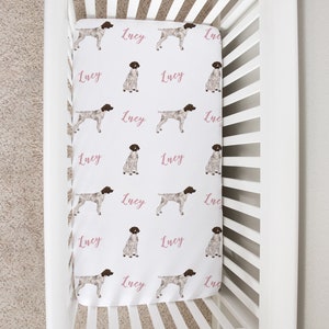 May include: White crib with a white fitted sheet featuring a repeating pattern of brown and white dogs and the name "Lucy" in pink lettering.