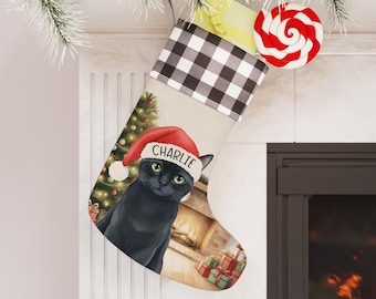Peut inclure: Une chaussette de Noël avec un revers à carreaux noir et blanc. La chaussette représente un chat noir portant un chapeau de Père Noël avec le nom "Charlie". Un sapin de Noël et des cadeaux sont également représentés.