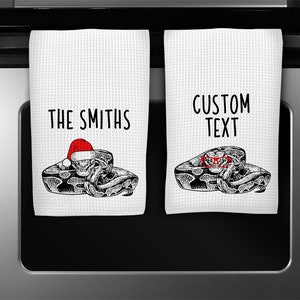 May include: Two white kitchen towels with a black and white illustration of a snake wearing a Santa hat and sunglasses. The text on the towels reads "THE SMITHS" and "CUSTOM TEXT".