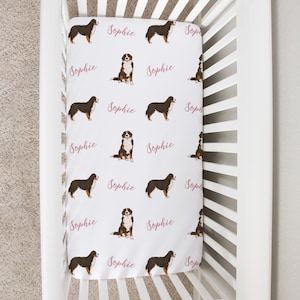 May include: White crib with a white and pink fitted sheet featuring a repeating pattern of Bernese Mountain Dogs and the name "Sophie".