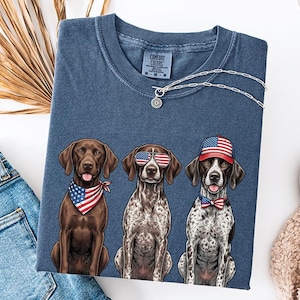May include: A blue Comfort Colors t-shirt featuring three German Shorthaired Pointers. Each dog is wearing patriotic accessories: a bandana, sunglasses, and a hat, all in red, white, and blue. A silver necklace with a pendant is also visible.