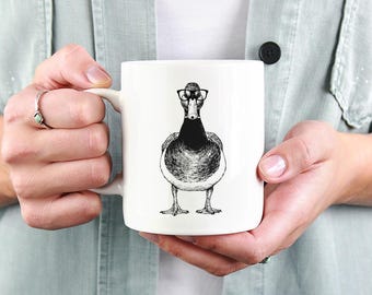 May include: White ceramic mug featuring a black and white illustration of a duck wearing glasses. The duck is centered on the mug, with a detailed drawing style. The mug has a standard handle and a smooth, glossy finish.