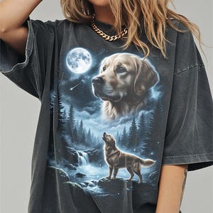 May include: Dark gray t-shirt featuring a detailed graphic of a golden retriever under a full moon in a forest setting. The design includes a waterfall and a howling dog, creating a serene, nature-inspired scene. The shirt has a vintage, washed look.