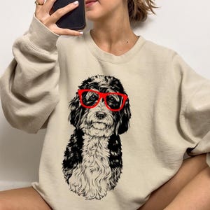 May include: A beige sweatshirt featuring a black and white illustration of a dog wearing red glasses. The dog has a curly coat and is looking directly at the viewer.
