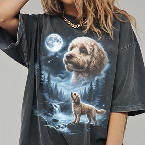 May include: Dark gray t-shirt featuring a detailed illustration of a dog in a forest scene under a full moon. The design includes a waterfall, trees, and a starry sky, creating a whimsical and nature-inspired aesthetic.