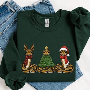 May include: A dark green sweatshirt featuring three cartoon snakes in festive holiday attire. One snake wears a reindeer antler, another a Christmas tree, and the third a Santa hat. The snakes are brown and tan.