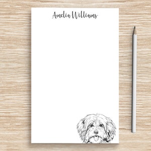 May include: A white notepad with the name "Amelia Williams" in script at the top, a black and white illustration of a dog at the bottom, and a white pencil on a wooden surface.
