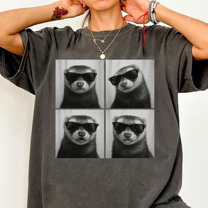 May include: Dark grey t-shirt featuring a four-panel image of a ferret wearing sunglasses. The ferret is in different poses in each panel. The shirt is a relaxed fit and has a vintage look.