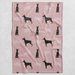 Puede incluir: Manta de forro polar rosa con un patr&oacute;n repetido de perros Doberman Pinscher negros y marrones y el nombre "Sophie" en texto blanco.
