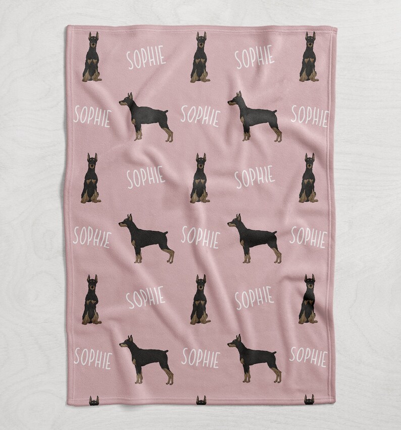 Puede incluir: Manta de forro polar rosa con un patr&oacute;n repetido de perros Doberman Pinscher negros y marrones y el nombre "Sophie" en texto blanco.