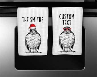 May include: Two white kitchen towels with a waffle weave pattern. One towel features the text "THE SMITHS" above a black and white bird illustration wearing a Santa hat. The other towel has "CUSTOM TEXT" above a bird with red glasses.