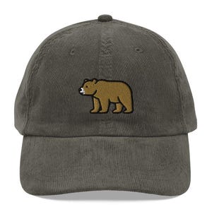 May include: A dark green corduroy baseball cap with an embroidered brown bear and white snout. The cap has a curved peak and a structured crown. This hat is a casual accessory.