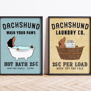 Puede incluir: Dos láminas enmarcadas con ilustraciones de perros salchicha. Una muestra un perro salchicha en una bañera con el texto "DACHSHUND WASH YOUR PAWS HOT BATH 25C". La otra muestra un perro salchicha en una cesta de lavandería con el texto "DACHSHUND LAUNDRY CO. 25C PER LOAD".