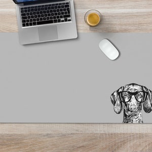 May include: A gray desk mat with a black and white illustration of a dog wearing glasses. The dog is looking to the right. There is a laptop, a coffee cup, and a computer mouse on the desk mat.