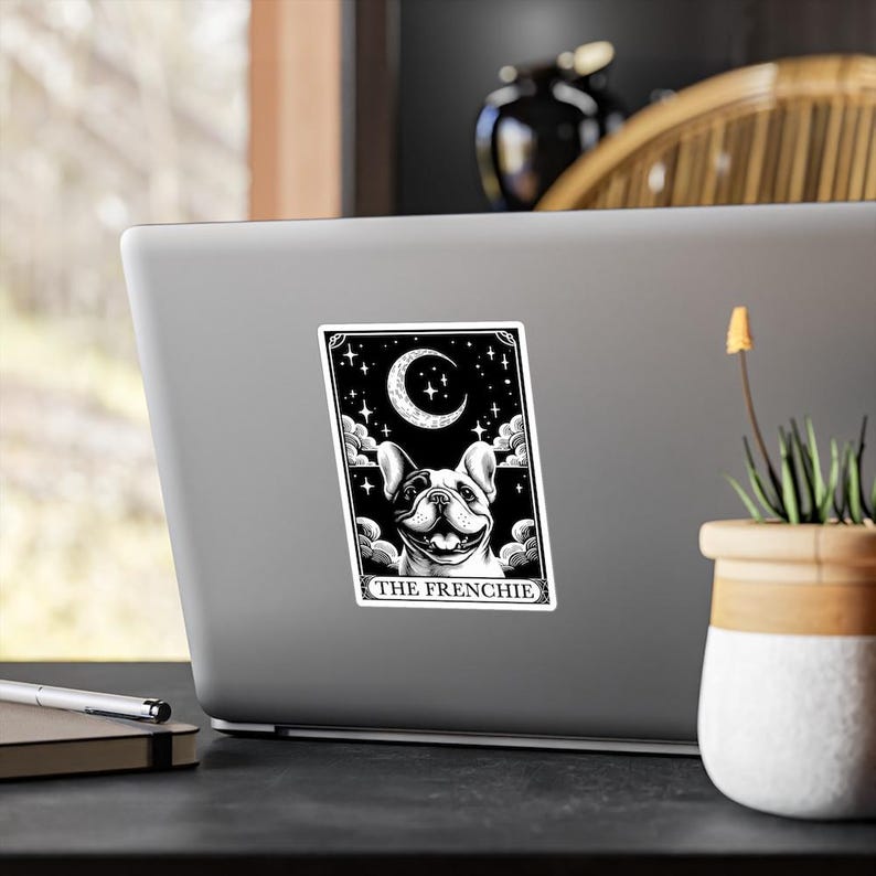 Op de afbeelding: Een zwart-witte sticker met een Franse Bulldog tarotkaart ontwerp. De kaart toont een lachende hond onder een halve maan en sterren, met de tekst "THE FRENCHIE" onderaan. De sticker zit op een laptop.