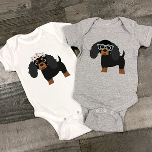 May include: Two white baby onesies with a black and brown Cavalier King Charles Spaniel dog graphic. One onesie has a floral crown on the dog and the other has blue glasses.