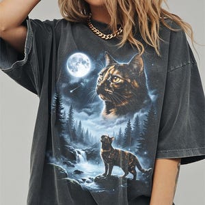 May include: Dark gray t-shirt featuring a detailed graphic of a cat under a full moon in a forest setting. The design includes a waterfall, trees, and a starry sky, creating a nature-inspired scene. The cat is depicted in two sizes, one large and one small.