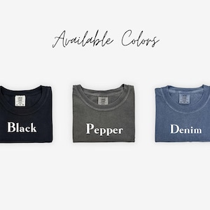 May include: Three folded t-shirts in different colors: black, pepper, and denim. The shirts are laid out on a white background with the word "Available Colors" written above them.