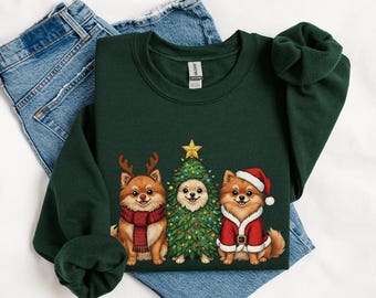 May include: A dark green sweatshirt featuring a festive design with three Pomeranian dogs. One dog wears a reindeer antler headband and a scarf, another is dressed as a Christmas tree, and the third wears a Santa hat and coat.