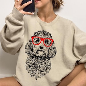 May include: A beige sweatshirt with a black and white illustration of a poodle wearing red glasses. The poodle is looking directly at the viewer.