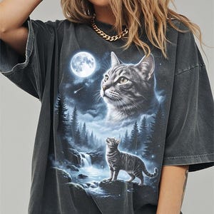 May include: Dark gray oversized t-shirt featuring a detailed graphic of a cat gazing at the moon in a forest scene. The design includes a waterfall, trees, and a second cat. The shirt has short sleeves and a relaxed fit.