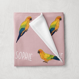 May include: A pink fleece blanket with a white fleece reverse side. The blanket features a repeating pattern of yellow, green, and blue parrots. The text "SOPHIE" is printed in white on the blanket.