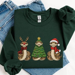 May include: A dark green sweatshirt with a festive design. It features three snakes, one with reindeer antlers and a scarf, another as a Christmas tree, and a third wearing a Santa hat and scarf.