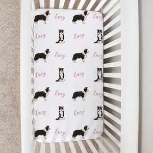 May include: White crib sheet with a repeating pattern of black and white border collie dogs and the name "Lucy" in pink lettering.