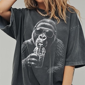 May include: A charcoal gray oversized t-shirt featuring a white graphic print of a gorilla wearing sunglasses and drinking from a clear plastic cup with a straw. The shirt has a faded, washed-out appearance with short sleeves and a crew neckline.