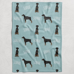 Puede incluir: Una manta de franela azul claro con un patr&oacute;n repetido de perros Doberman Pinscher negros y el nombre "Sophie" impreso en blanco.