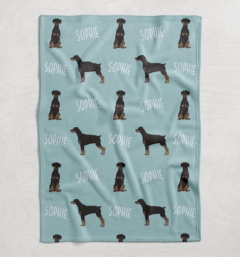 Puede incluir: Una manta de franela azul claro con un patr&oacute;n repetido de perros Doberman Pinscher negros y el nombre "Sophie" impreso en blanco.