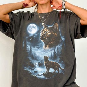 May include: Dark gray t-shirt featuring a detailed graphic of a cat under a full moon, with a waterfall and forest scene. The cat is depicted in two sizes, one large and one smaller, with a starry sky background. The shirt has short sleeves.
