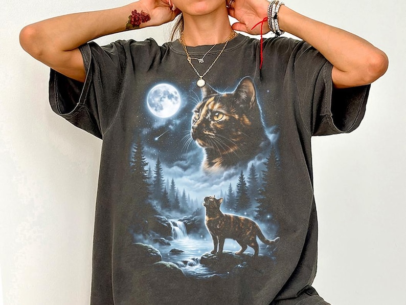May include: Dark gray t-shirt featuring a detailed graphic of a cat under a full moon, with a waterfall and forest scene. The cat is depicted in two sizes, one large and one smaller, with a starry sky background. The shirt has short sleeves.