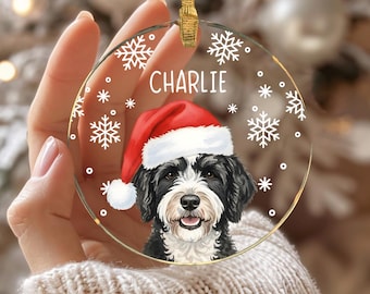 May include: A round, clear acrylic Christmas ornament featuring a printed illustration of a black and white dog wearing a red and white Santa hat. The name CHARLIE is printed in white above the dog, surrounded by white snowflake patterns.
