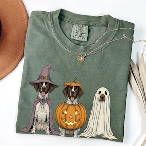 May include: A sage green short-sleeve t-shirt featuring a graphic of three dogs in Halloween costumes: one wearing a purple witch hat and cape, one inside a carved pumpkin, and one draped in a white ghost sheet. The shirt has a Comfort Colors label.