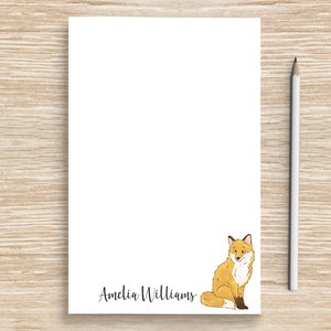 May include: A white notepad with a fox illustration and the name "Amelia Williams" printed at the bottom. The notepad is on a woodgrain surface with a pencil beside it.