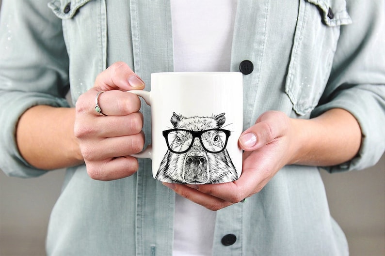 May include: White ceramic mug with a black and white illustration of a capybara wearing glasses. The capybara is looking directly at the viewer.