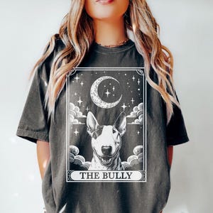May include: A grey t-shirt featuring a white graphic of a bull terrier dog in a tarot card style design. The card reads "The Bully" and includes a crescent moon and stars.