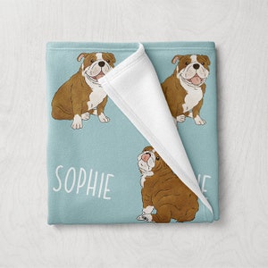 May include: A blue fleece blanket with a white fleece lining. The blanket features a repeated pattern of brown and white bulldogs with the name "Sophie" printed on the blanket.