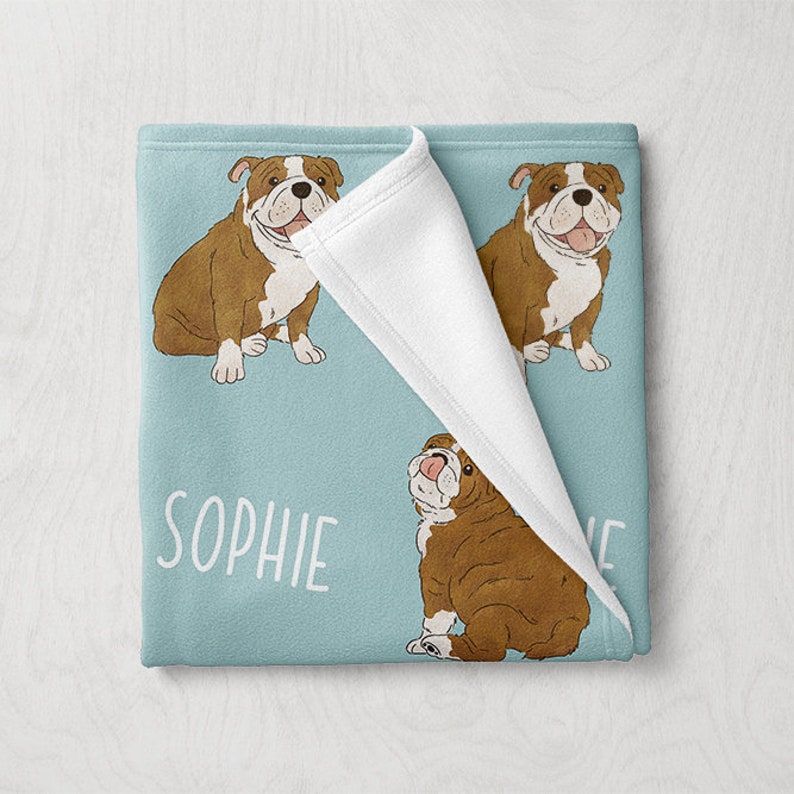 May include: A blue fleece blanket with a white fleece lining. The blanket features a repeated pattern of brown and white bulldogs with the name "Sophie" printed on the blanket.