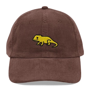 Peut inclure: Casquette de baseball en velours côtelé marron avec un motif de lézard brodé en jaune. La casquette a une visière incurvée et une sangle réglable. Un accessoire décontracté pour un usage quotidien.